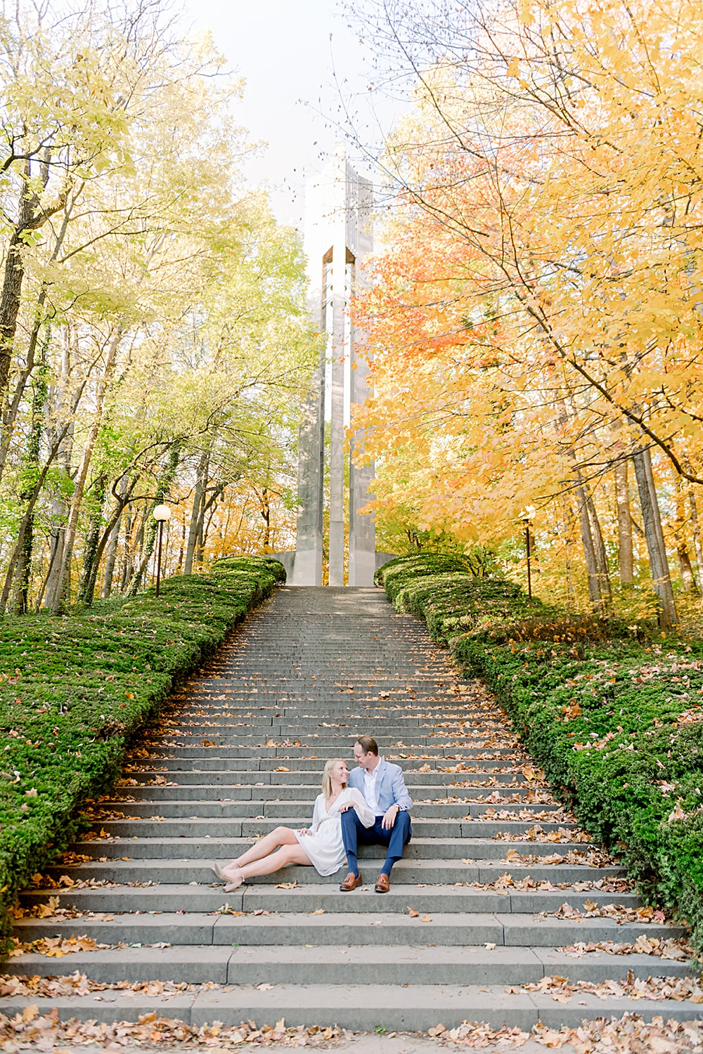 A couple sits on the staircase in front of the clock tower at Butler University. Stunning fall foliage surrounds them at Holcomb Gardens.