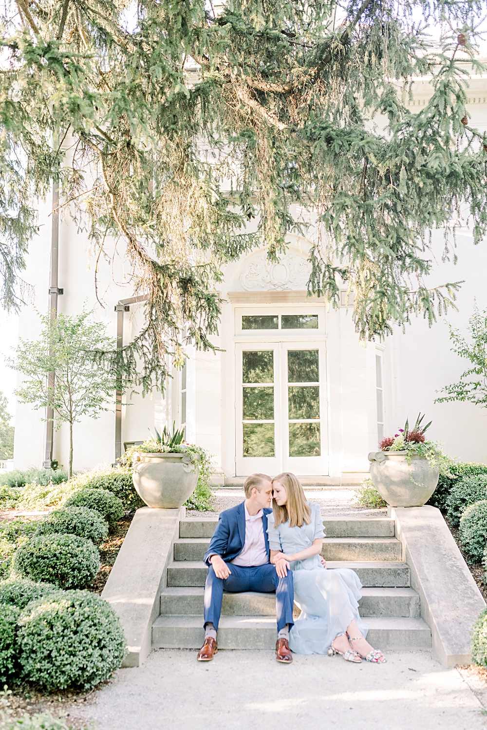 Couple sitting on cement steps under a large pine tree. Golden light is falling through the branches creating a beautiful glow around them. She is in a blue dress and he is in a blue suit.