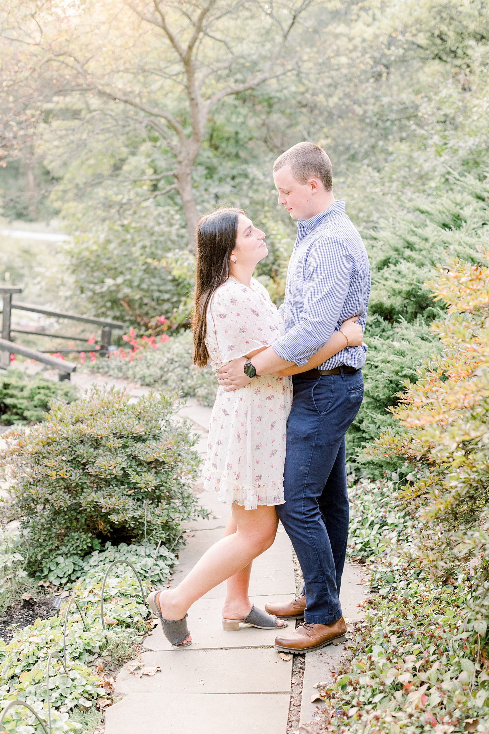 Couple embracing on garden path at Newfields in Indianapolis. The woman is wearing a short white dress and the man is in a blue dress shirt.