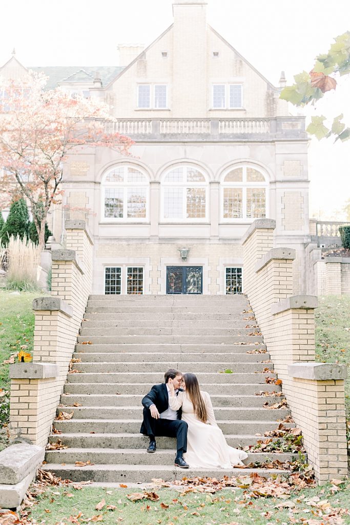 A couple is sitting on the steps out side of Laurel Hall in Indianapolis, Indiana during their engagement session. There are fallen leaves all around them and you can see the tan building behind them.