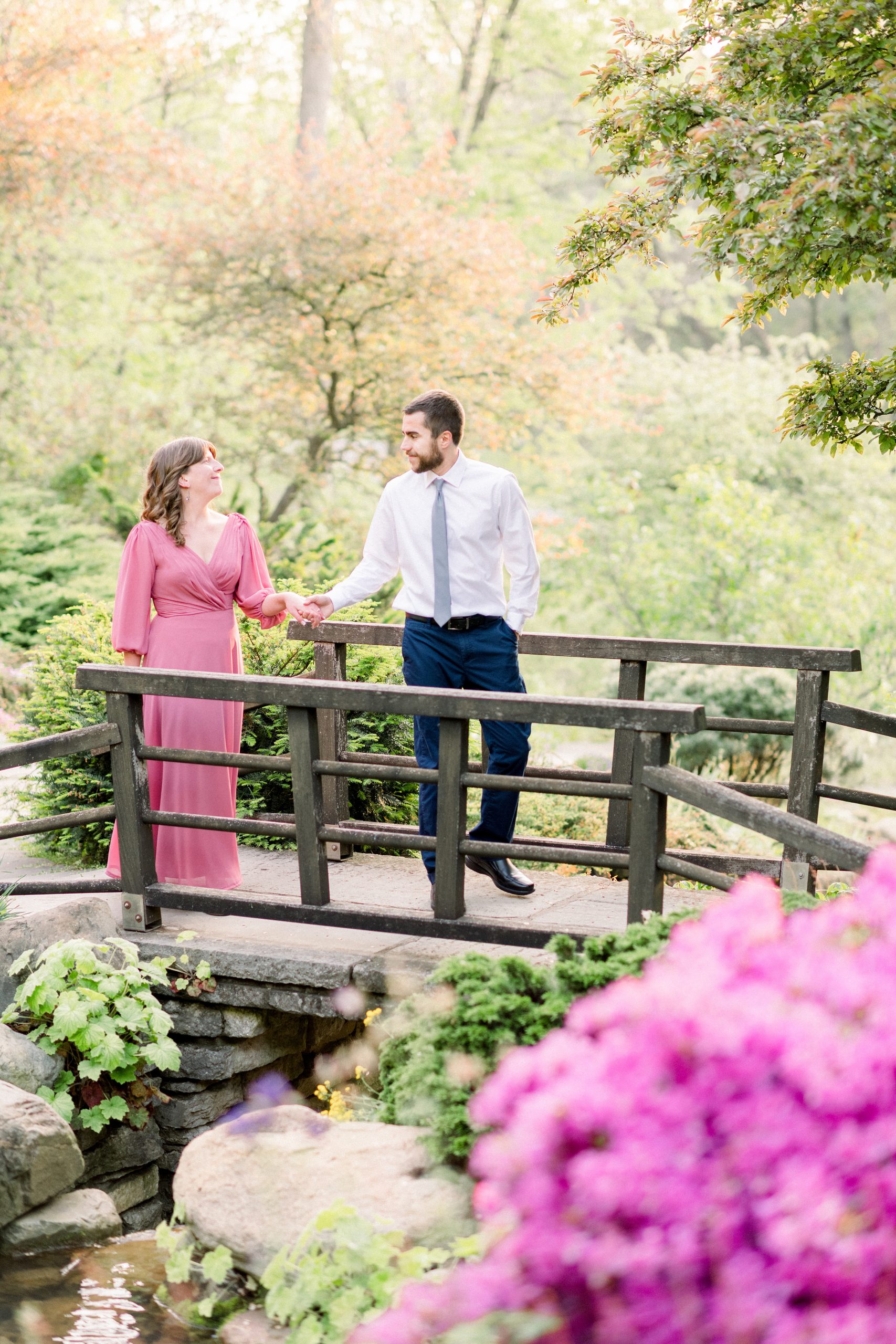 Woman in pink dress is escorted over bridge by man in white dress shirt at Newfields in Indianapolis during their engagement photos