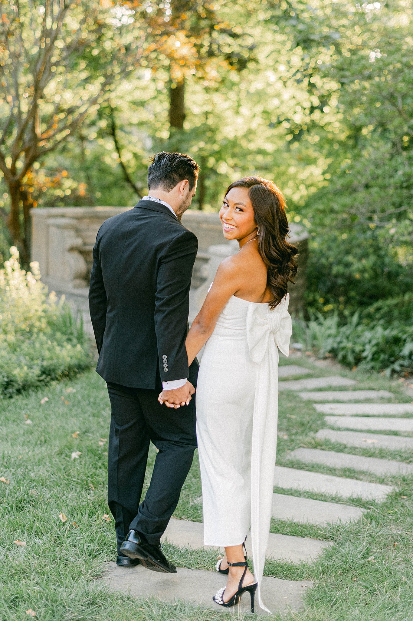 A couple walks away from the camera holding hands. They are strolling down a path into a garden at Newfields. The man is wearing a black suit and is looking towards the path. She is wearing a white dress with a large white bow on the pack and looking back over her shoulder.