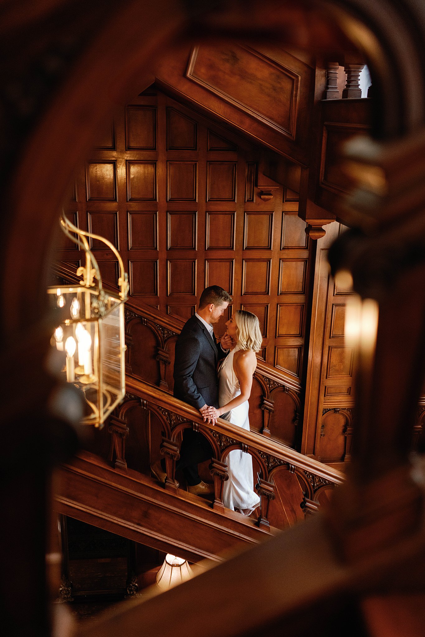 A couple steal a kiss on the staircase of Laurel Hall. He is standing on the step above her and her hand is resting on the wooden hand railing.