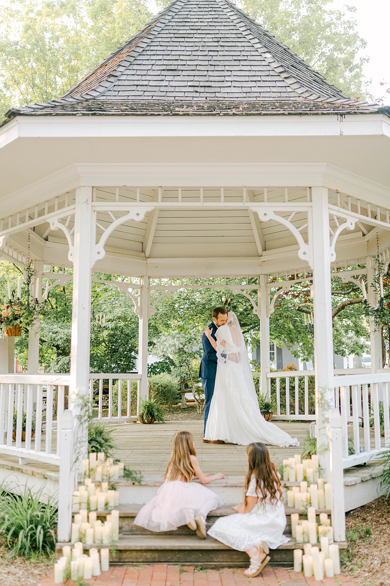 Bride and groom share their first dance under the gazebo at a park in Zionsville, Indiana. Two little girls sit on the steps of the gazebo watching.