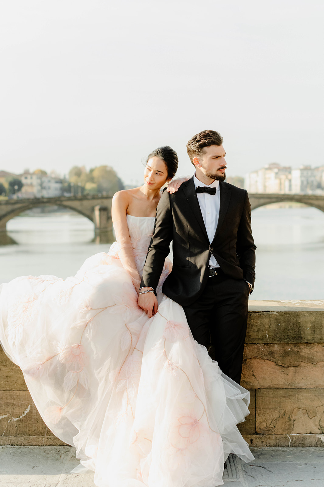 Bride and groom sitting together on a stone ledge overlooking the Arno River