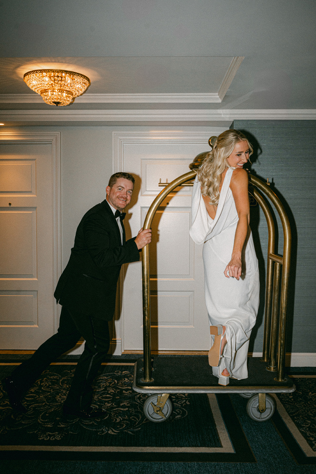 Groom playfully pushes bride on a gold luggage cart at Hotel Carmichael, bride smiling in a sleek white gown with open back