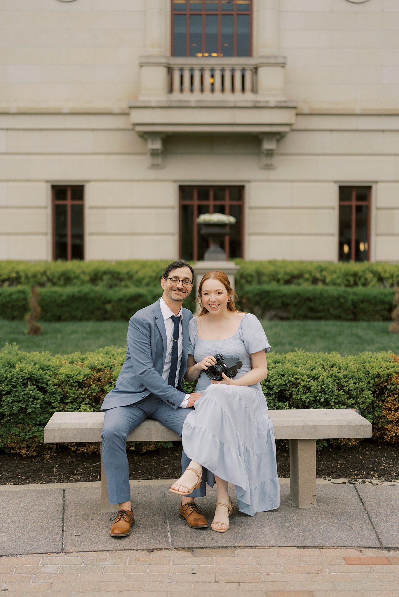 Alison and Zach of Alison Mae Photography sitting on a bench in front of a large historic building